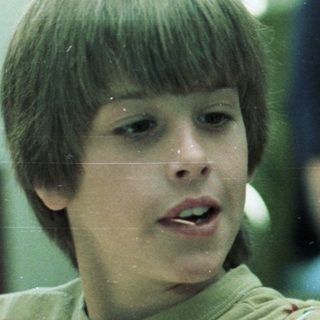 Young boy playing video game with adult in background.