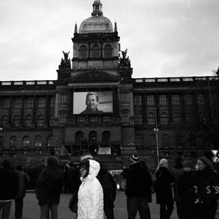 People gather in front of Prague Castle displaying a large portrait.