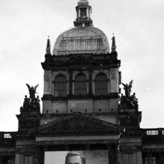 People gather in front of Prague Castle displaying a large portrait.