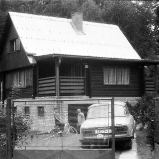Rural wooden house with parked car and man gardening.