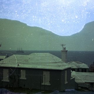Coastal town with houses, hills, and distant ships.