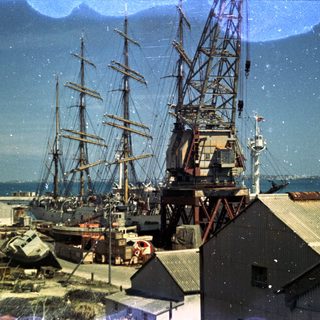 Historic sailing ships docked near industrial cranes.