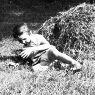 Woman standing beside haystack, child lying on grass.