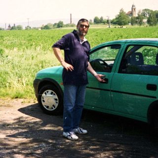 Man standing beside open hatch of green car in rural field.
