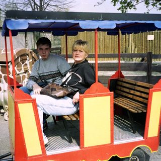 Two people riding a small, open train cart at zoo.