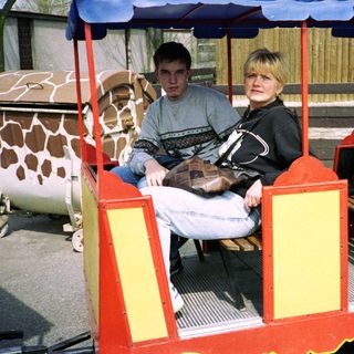 Two people riding a small, open train cart at zoo.