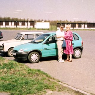 Two people standing by a turquoise car in a parking lot.