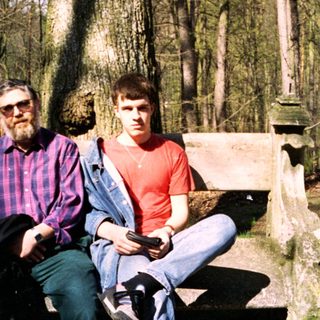 Two men sitting on a bench in a wooded cemetery.