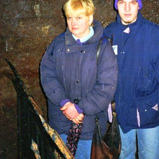 Two people standing indoors near a dark stone wall.