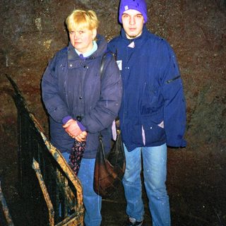 Two people standing indoors near a dark stone wall.
