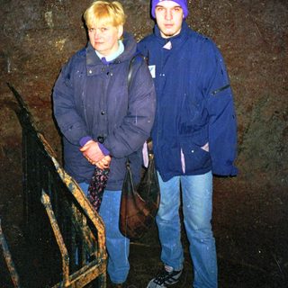Two people standing indoors near a dark stone wall.