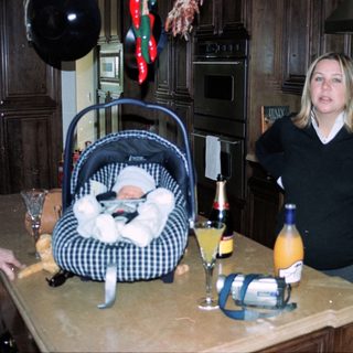 Newborn in carrier, woman standing by kitchen counter with drinks.