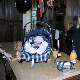 New mother posing with baby in carrier, celebrating with drinks on kitchen counter.