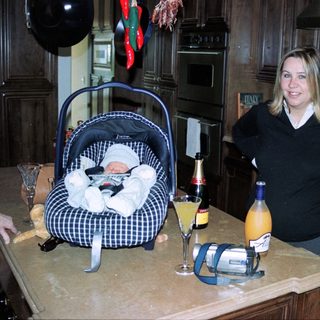 New mother posing with baby in carrier, celebrating with drinks on kitchen counter.