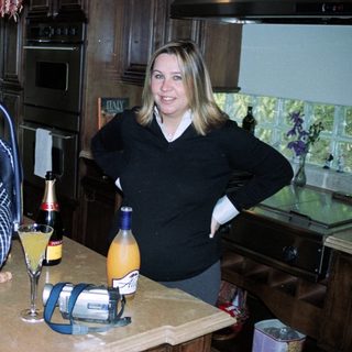 New mother posing with baby in carrier, celebrating with drinks on kitchen counter.
