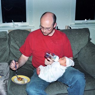 Man holding baby while eating pizza on couch.