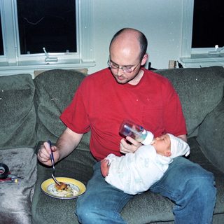 Man holding baby while eating pizza on couch.