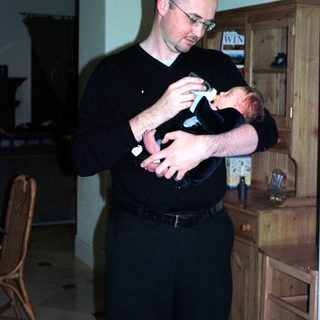 Man holding baby indoors near wooden cabinet.
