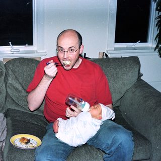 Man eating while holding baby on couch.