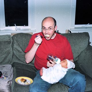 Man eating while holding baby on couch.