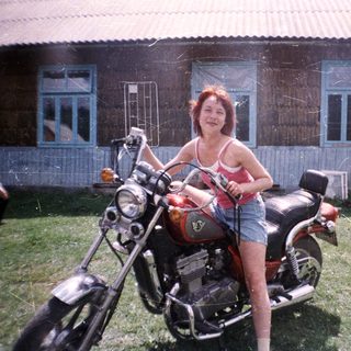 Woman posing on classic motorcycle outside rustic house.