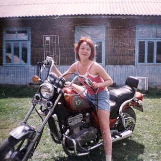 Woman posing on classic motorcycle outside rustic house.