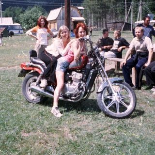 Two women posing on vintage motorcycle at outdoor gathering.
