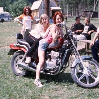 Two women posing on vintage motorcycle at outdoor gathering.
