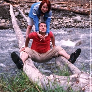 Two people sitting on fallen log near river.