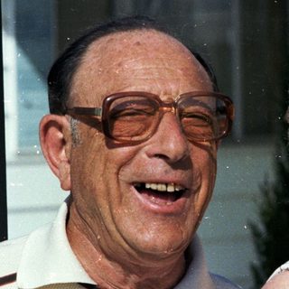 Two elderly couple smiling outdoors near window.
