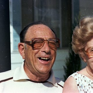 Two elderly couple smiling outdoors near window.