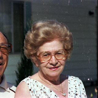Two elderly couple smiling outdoors near window.