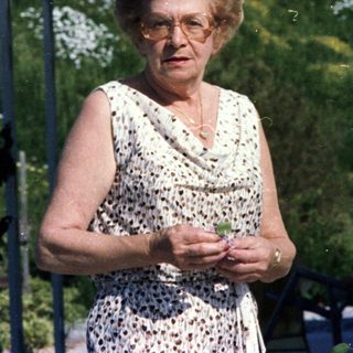 Elderly woman standing outdoors near house.