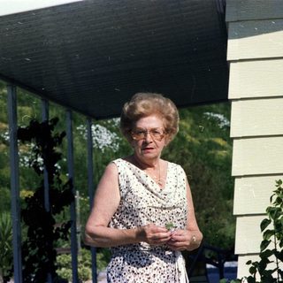 Elderly woman standing outdoors near house.