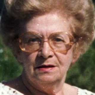 Elderly woman standing outdoors near house.