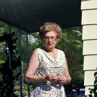 Elderly woman standing outdoors near house.