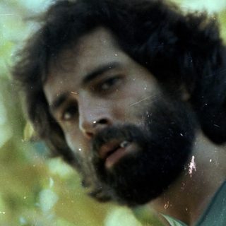 Man with beard standing outdoors among trees.
