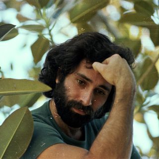 Man resting hand on forehead amidst green foliage.