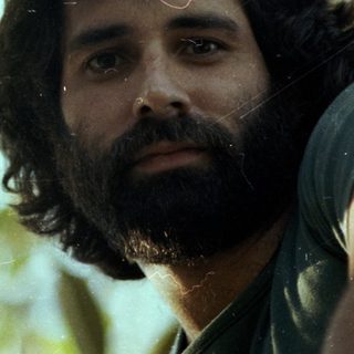 Man with beard outdoors among green foliage.