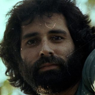 Man with beard outdoors among green foliage.