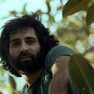 Man with beard outdoors among green foliage.