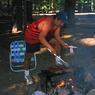 Man grilling meat over campfire outdoors.