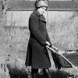 A woman in vintage clothing pushes an old-fashioned baby carriage outdoors.