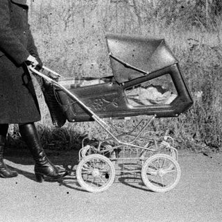 A woman in vintage clothing pushes an old-fashioned baby carriage outdoors.