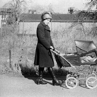 A woman in vintage clothing pushes an old-fashioned baby carriage outdoors.