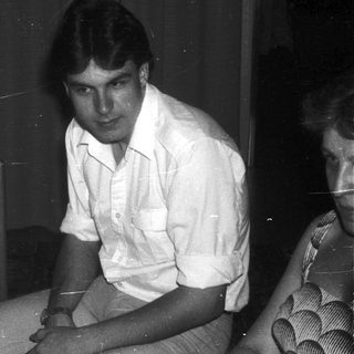 Two people sitting indoors in casual 1970s-style clothing.