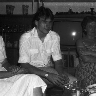 Two people seated indoors at a table, dressed in 1970s-80s style.