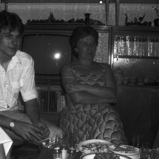 Two people seated indoors at a table, dressed in 1970s-80s style.