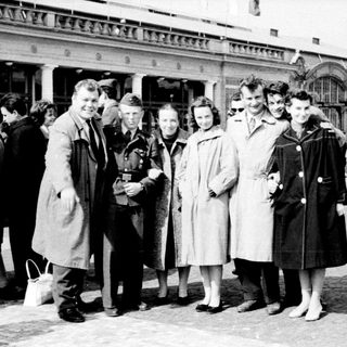 Group of people posing outside a train station in mid-20th century attire.