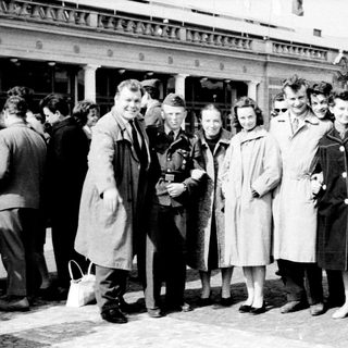 Group of people posing outside a train station in mid-20th century attire.